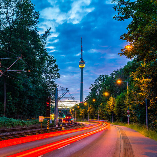 Lichtstreifen von Autos in der Dämmerung auf einer Straße. Im Hintergrund sieht man den Fernsehturm.