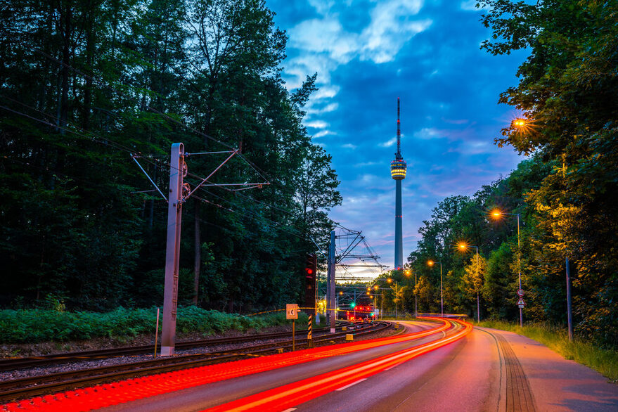 Lichtstreifen von Autos in der Dämmerung auf einer Straße. Im Hintergrund sieht man den Fernsehturm.