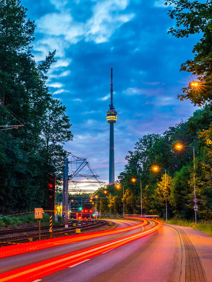 Lichtstreifen von Autos in der Dämmerung auf einer Straße. Im Hintergrund sieht man den Fernsehturm.