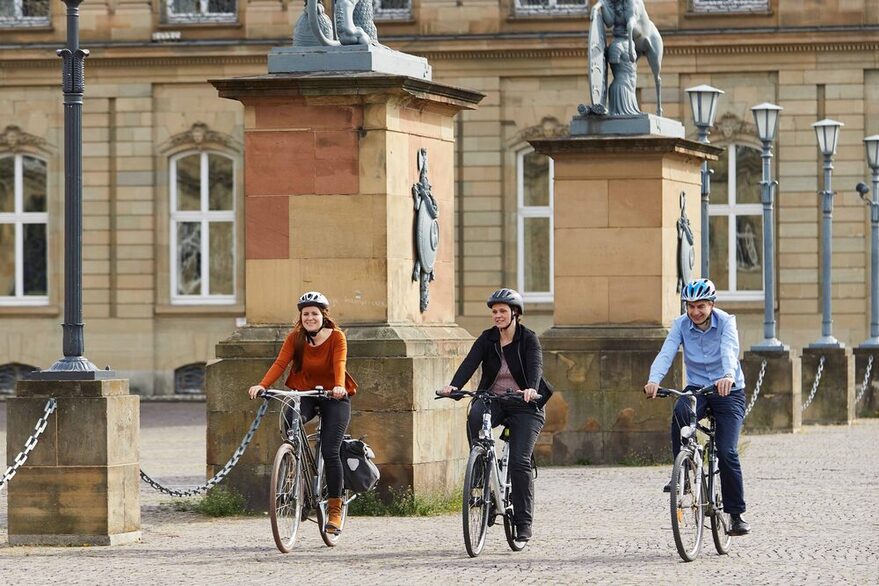 Drei Menschen radeln an einem Schloss vorbei.