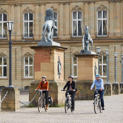 Drei Menschen radeln an einem Schloss vorbei.