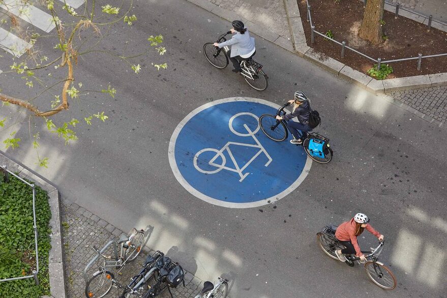 Drei Fahrradfahrer fahren auf einer Fahrradstraße, erkennbar an einem großen, blauen Zeichen auf dem Asphalt.