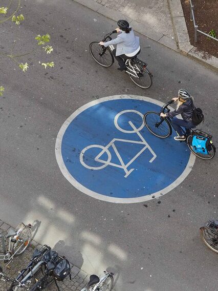 Drei Fahrradfahrer fahren auf einer Fahrradstraße, erkennbar an einem großen, blauen Zeichen auf dem Asphalt.