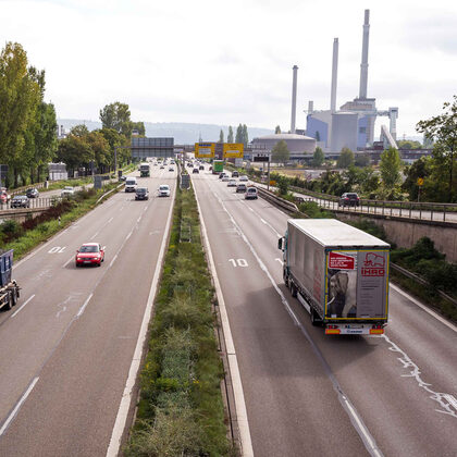 Blick auf eine Bundesstraße mit vielen Autos und Lastwagen. Links im Hintergrund ein große Fabrikhalle.