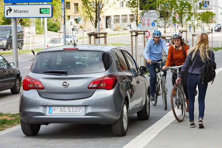 Zwei Radfahrer überholen ein parkendes Auto und schneiden eine Fußgängerin.