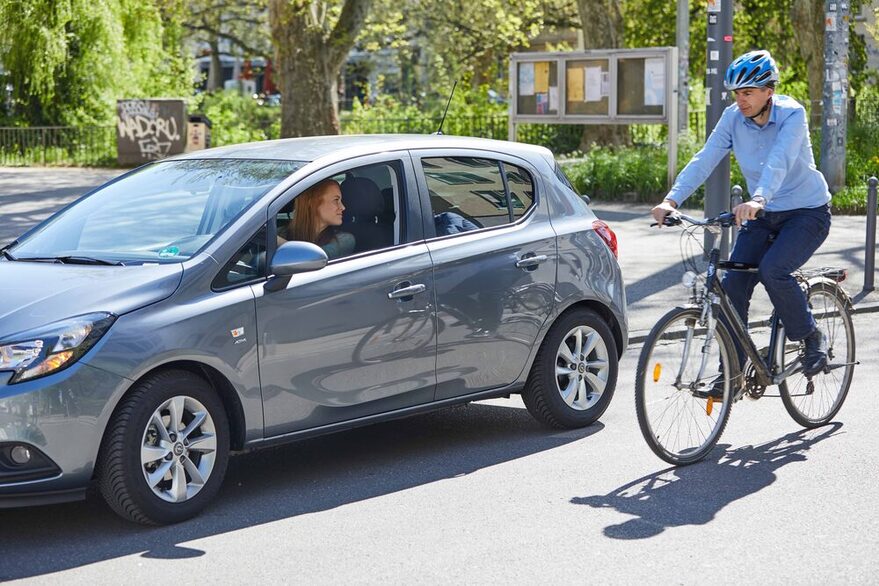 Eine Frau im Auto macht einen Schulterblick, ein Fahrradfahrer fährt vorbei.