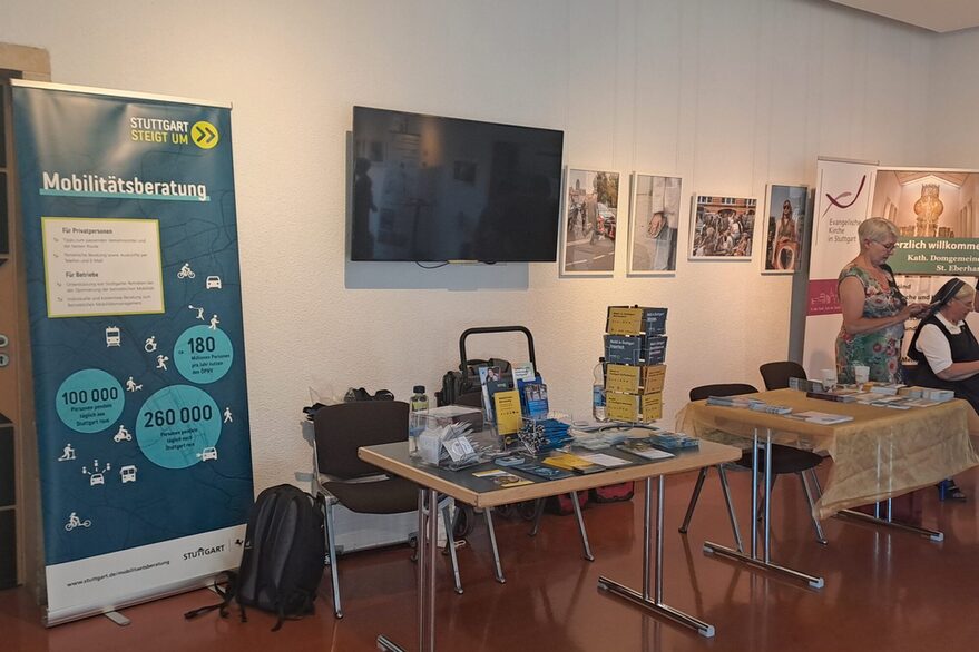 Es ist der Stand der Mobilitätsberatung zu sehen: ein großer Tisch mit mehreren Flyer-Ständern, auf dem auch diverse Mobilitätskarten ausgelegt sind. Schräg links hinter dem Stand ist ein Roll-up zu sehen, auf dem die Mobilitätsberatung beworben wird. Rechts neben dem Tisch ist ein weiterer Tisch eines anderen Anbieters zu sehen.