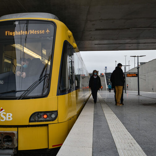 Eine Straßenbahn der SSB steht an der Haltestelle Flughafen/messe, während einige Menschen auf dem Bahnsteig stehen.