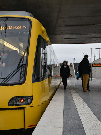 Eine Straßenbahn der SSB steht an der Haltestelle Flughafen/messe, während einige Menschen auf dem Bahnsteig stehen.