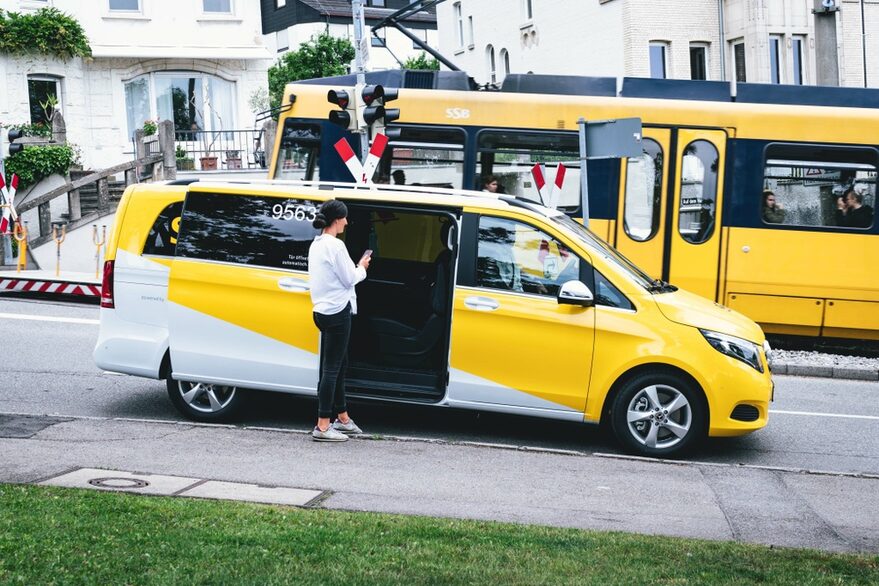 Eine Frau steht vor einem gelben Sprinter, im Hintergrund fährt eine gelbe Stadtbahn vorbei.