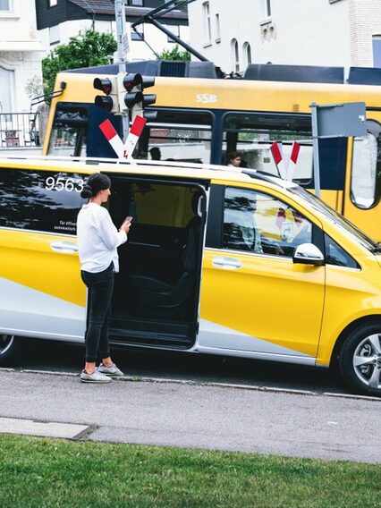 Eine Frau steht vor einem gelben Sprinter, im Hintergrund fährt eine gelbe Stadtbahn vorbei.