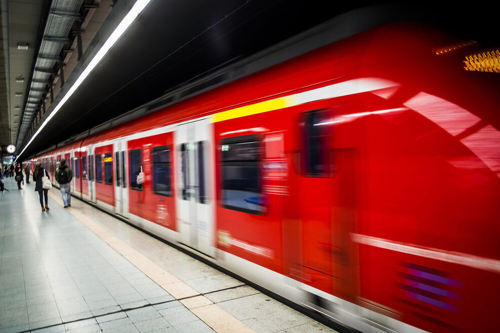 Eine rote S-Bahn fährt von einer Haltestelle ab. Am Gleis stehen Menschen.