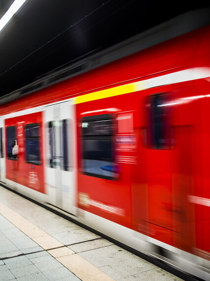 Eine rote S-Bahn fährt von einer Haltestelle ab. Am Gleis stehen Menschen.