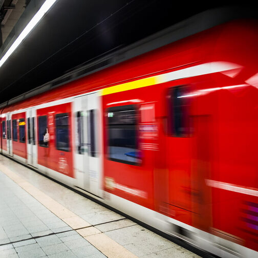 Eine rote S-Bahn fährt von einer Haltestelle ab. Am Gleis stehen Menschen.