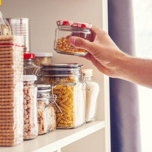 Vorratskammer: Eine Hand greift in ein Regal mit Gläsern, in denen sich Nudeln, Nüsse und andere Lebensmittel befinden.