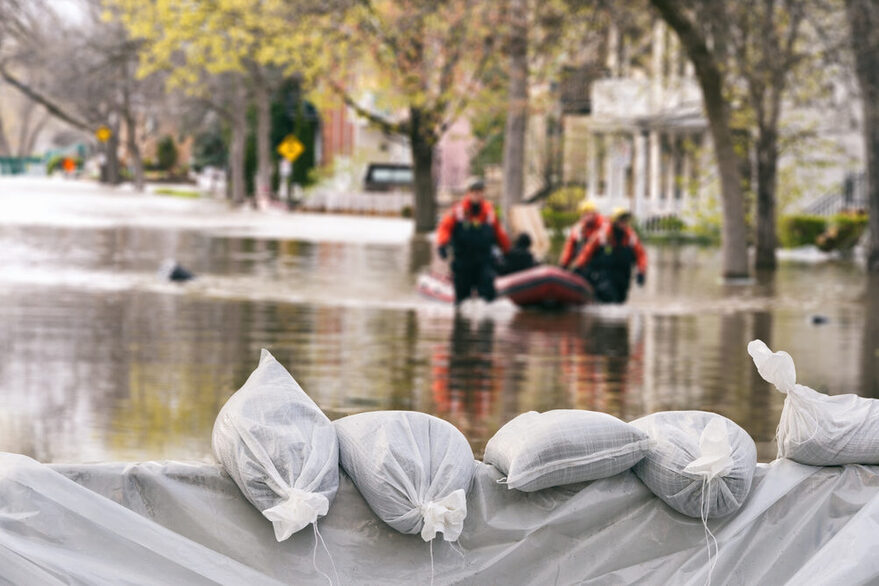 Eine überflutete Straße wird mit Sandsäcken gesichert. Rettungskräfte bergen Menschen mit einem Boot.