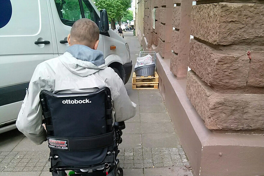 Ein rücksichtsloser Fahrer blockiert mit seinem weißen Transporter den Weg für einen Rollstuhlfahrer.