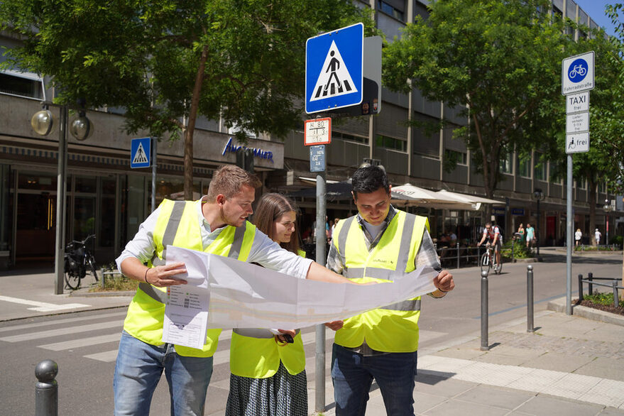 Städtische Mitarbeitende in gelben Sicherheitswesten stehen mit einer Karter in einer Fahrradtstraße