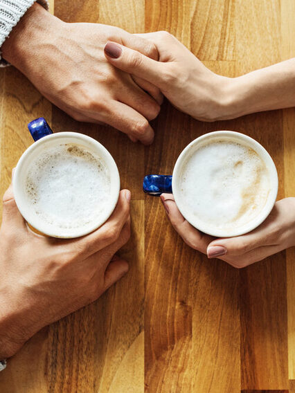 Situation am Esstisch: Zwei Menschen halten jeweils eine Kaffetasse in der Hand und berührern sich mit den Händen.