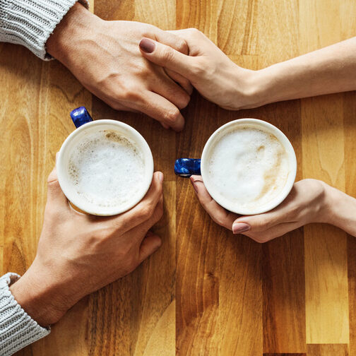 Situation am Esstisch: Zwei Menschen halten jeweils eine Kaffetasse in der Hand und berührern sich mit den Händen.