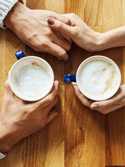 Situation am Esstisch: Zwei Menschen halten jeweils eine Kaffetasse in der Hand und berührern sich mit den Händen.