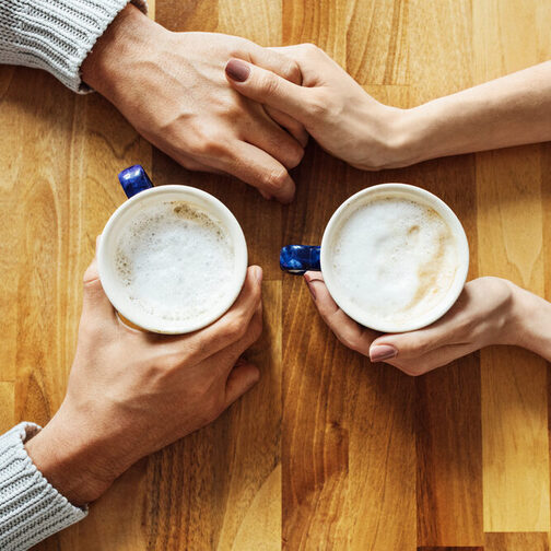 Situation am Esstisch: Zwei Menschen halten jeweils eine Kaffetasse in der Hand und berührern sich mit den Händen.