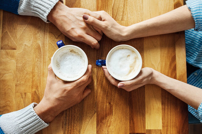 Situation am Esstisch: Zwei Menschen halten jeweils eine Kaffetasse in der Hand und berührern sich mit den Händen.