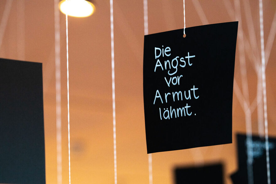 Schwarze Papierschilder hängen von der Decke. Ein Schild hat die Aufschrift: "Die Angst vor Armut lähmt."