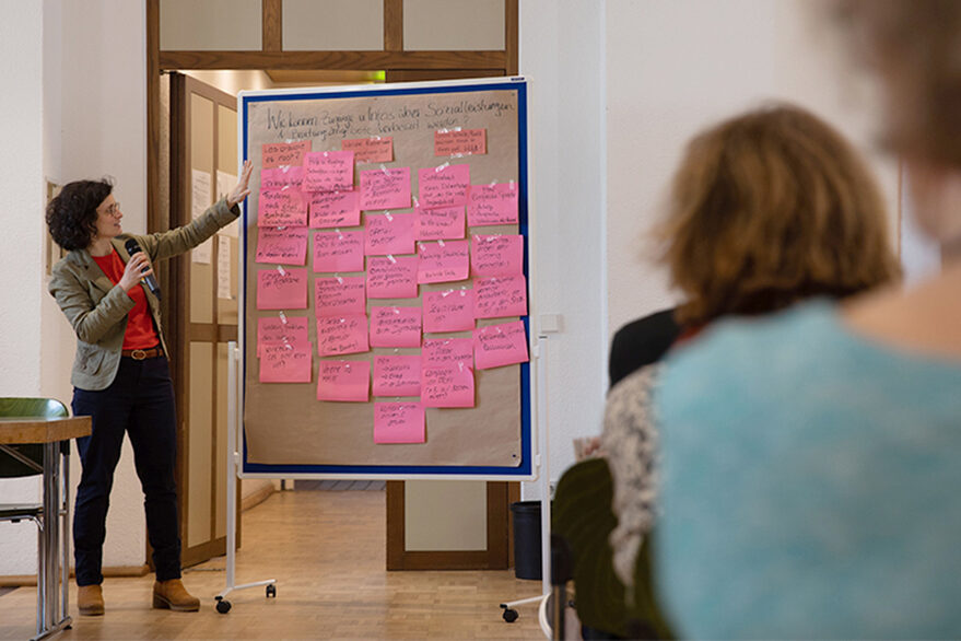 A woman with a microphone in her hand stands in front of a bulletin board with labeled index cards hanging on it. An audience listens to the woman.