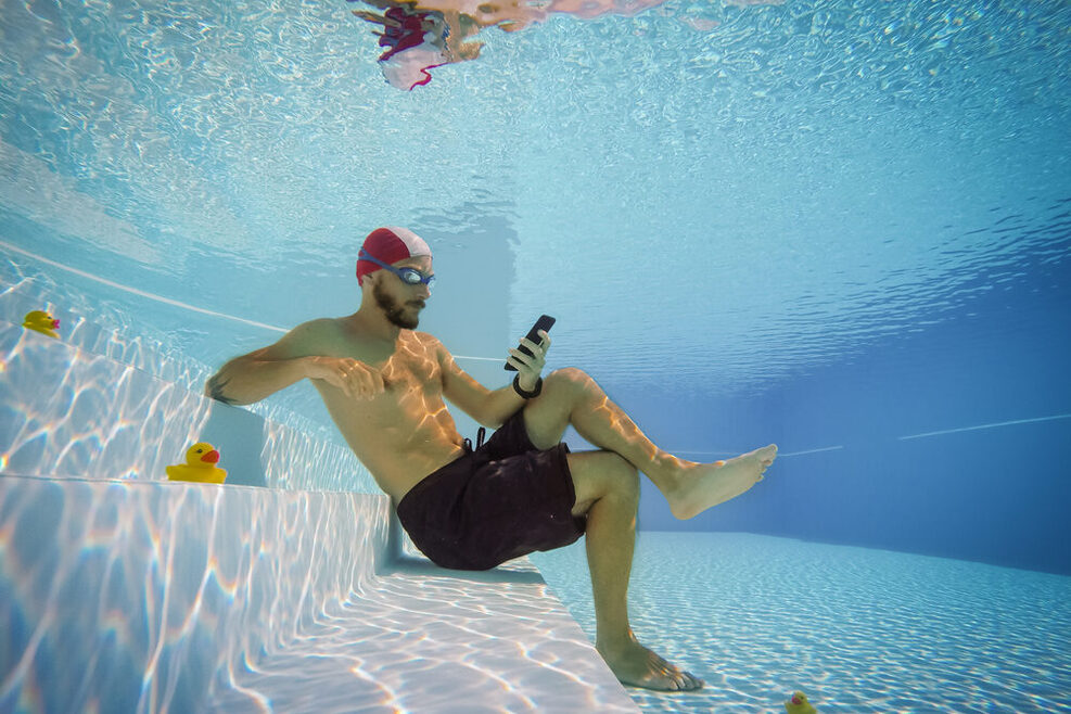 Ein Mann sitzt unter Wasser in einem Schwimmbad und liest auf seinem Smartphone.