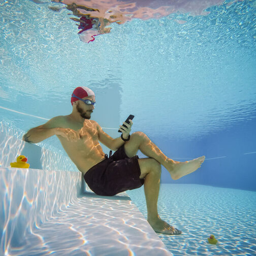 Ein Mann sitzt unter Wasser in einem Schwimmbad und liest auf seinem Smartphone.