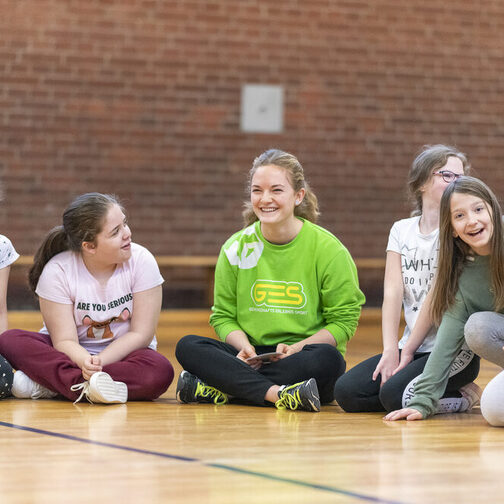 Eine Gruppe Kinder und eine Betreuerin sitzen auf dem Sporthallenboden.