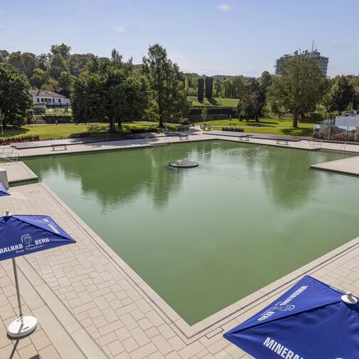 Schwimmbecken mit Wasser gefüllt. Sonnenschirme am Rand. Im Hintergrund Bäume mit Liegewiese.