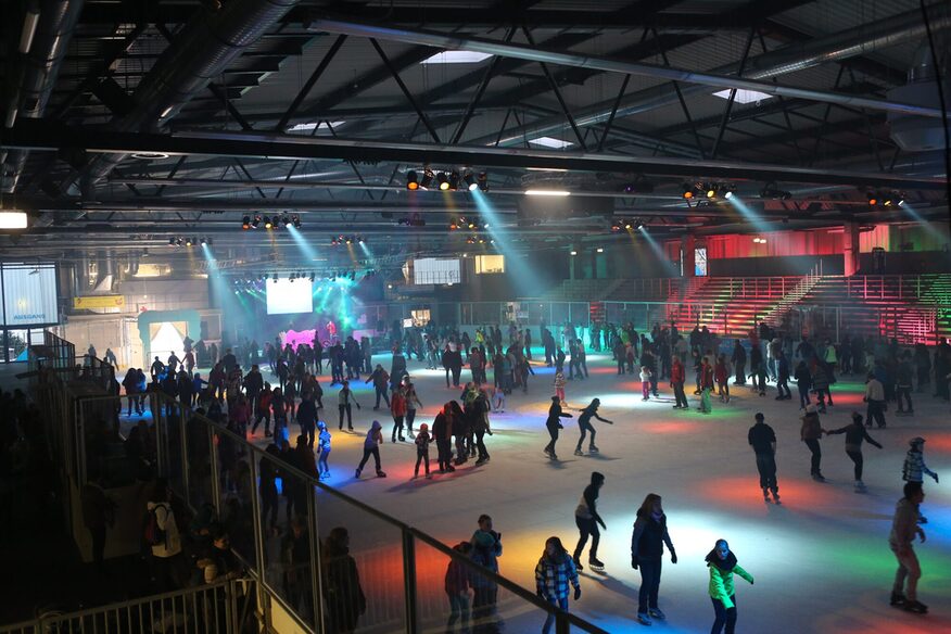 Eislaufhalle mit Besuchern auf der Eisfläche. Bunte Lichter erhellen den Raum.