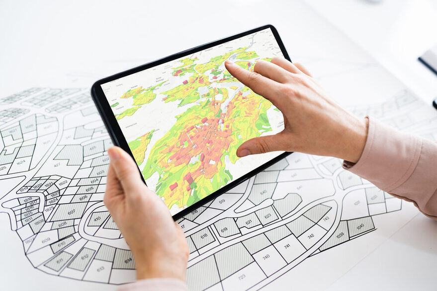Zu sehen ist ein Stadtplan, der auf einem Tisch liegt, während darüber Hände zu sehen sind, die ein teablet bedienen.