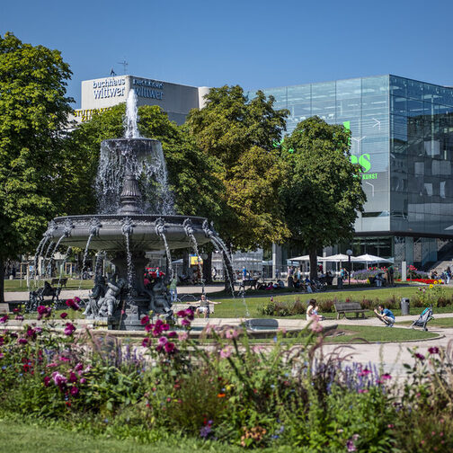 Blick über einen großen Platz in der Stadt mit viel Grünfläche und Bäumen. In der Mitte steht ein Brunnen.