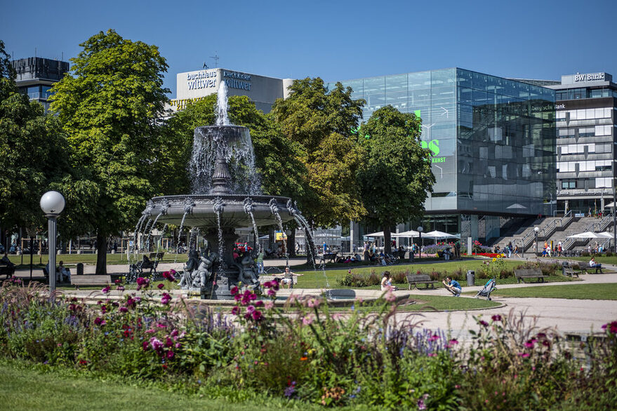 Blick über einen großen Platz in der Stadt mit viel Grünfläche und Bäumen. In der Mitte steht ein Brunnen.