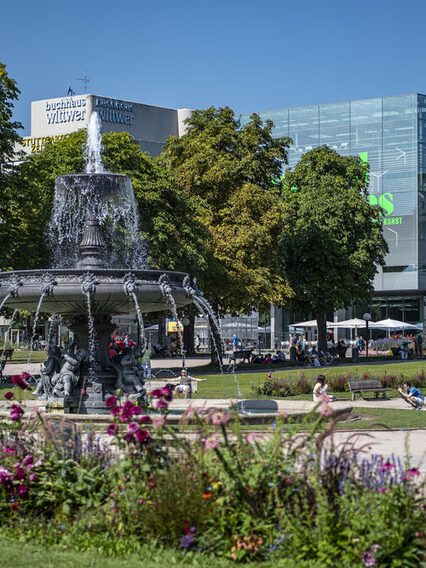 Blick über einen großen Platz in der Stadt mit viel Grünfläche und Bäumen. In der Mitte steht ein Brunnen.