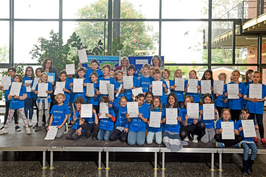 Mehrere Kinder in blauen T-Shirts sind auf eine Bühne und halten eine Urkunde vor sich.