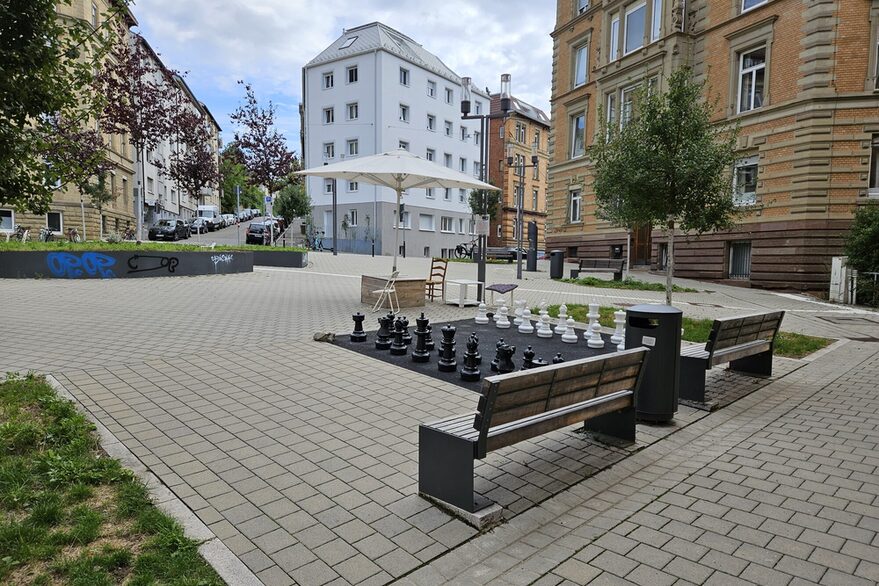 Der moderne Schützenplatz in Stuttgart mit zwei Sitzbänken, einem großen Schachbrett und einem Sonnenschirm. Im Hintergrund sieht man Bäume und Mehrfamilienhäuser.
