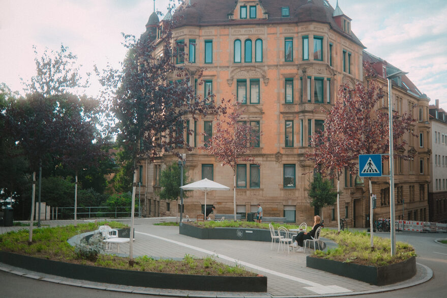 Blick auf ein Rondell am Schützenplatz mit Grünflächen, jungen Bäumen, Stühlen, auf denen eine Frau sitzt, und einem Sonnenschirm. Im Hintergrund ist ein Mehrfamilienhaus im Gründerstil zu sehen.
