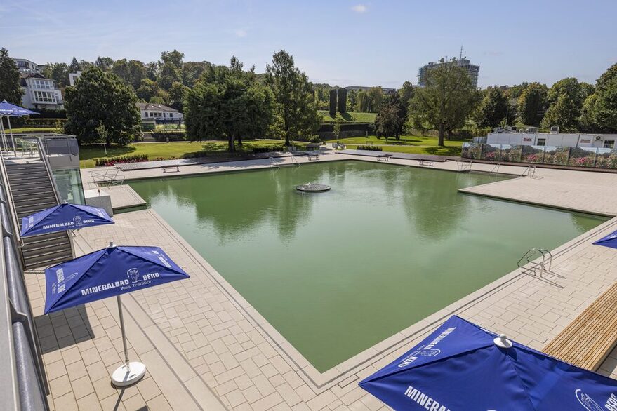 Schwimmbecken mit Wasser gefüllt. Sonnenschirme am Rand. Im Hintergrund Bäume mit Liegewiese.