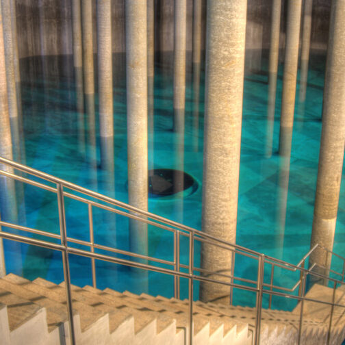 Innenansicht Trinkwasser-Hochbehälter. Eine lange und schmale Treppe führt in ein großes Wasserbecken in dem sich einige Steinsäulen befinden.