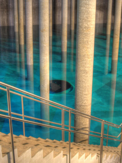 Innenansicht Trinkwasser-Hochbehälter. Eine lange und schmale Treppe führt in ein großes Wasserbecken in dem sich einige Steinsäulen befinden.