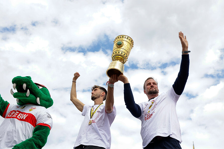 Das Foto zeigt VfB-Maskottchen Fritzle, Kapitän Atakan Karazor und Trainer Sebastian Hoeneß (von links nach rechts) mit dem DFB-Pokal auf der Tribüne auf dem Stuttgarter Schlossplatz, wie sie ihren Fans dankbar und ergriffen winken. Foto: Leif Piechowski/Stadt Stuttgart