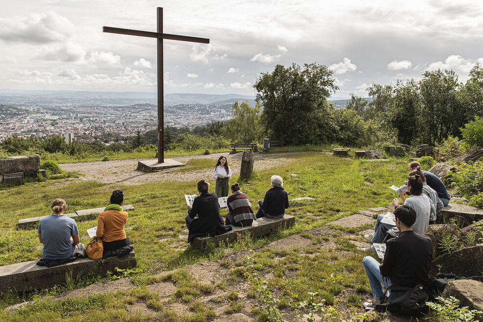 Mehrere Personen sitzen auf dem Birkenkopf in Stuttgart. Im Hintergrund thront ein großes Kreuz. In der Ferne ist im Tal die Landeshauptstadt Stuttgart zu sehen.