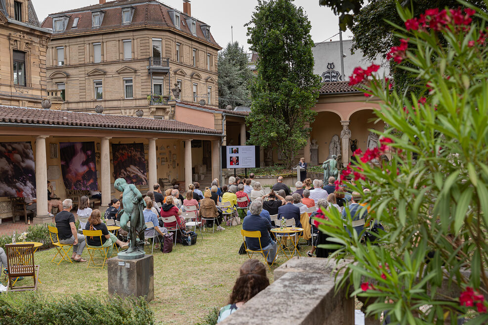 Mehrere Menschen sitzen auf Stühlen auf der Wiese im Städtischen Lapidarium in Stuttgart und hören einem Vortrag zu. Im Vordergrund ist ein blühender Oleander zu sehen, im Hintergrund ein Haus aus der Gründerzeit.