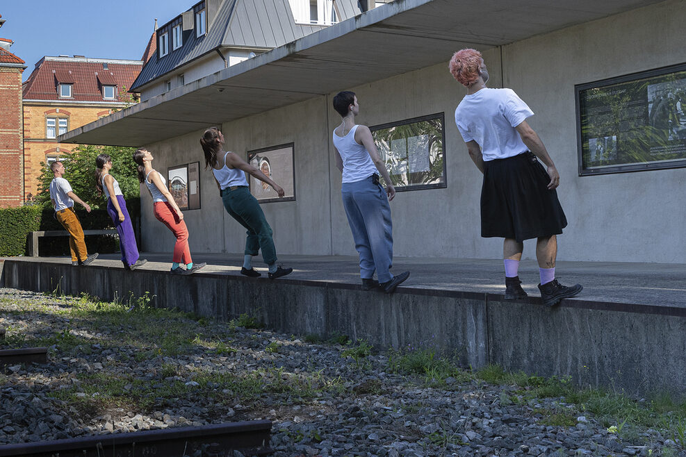 Sechs Personen in einer Reihe auf einer kleinen Mauer mit dem Rücken zur Kamera lehnen sich nach hinten, als würden sie rückwärts von der Mauer kippen.
