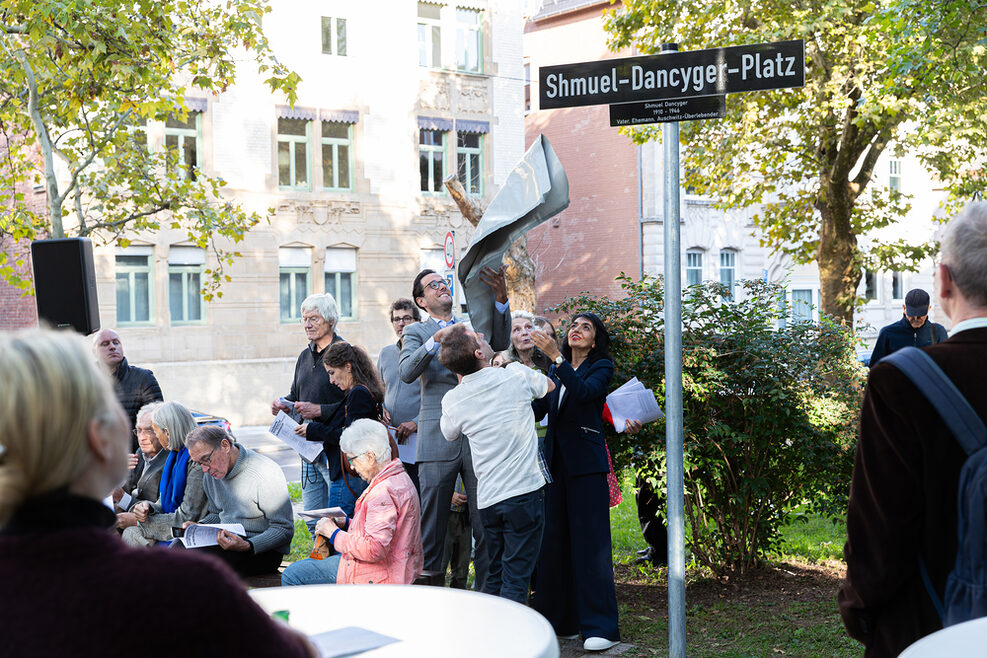 Erster Bürgermeister Dr. Fabian Mayer, der Enkel von Shmuel Dancyger und Landtagspräsidentin Muhterem Aras enthüllen auf einem Platz in Stuttgart-West das neue Straßenschild „Shmuel-Dancyger-Platz“.