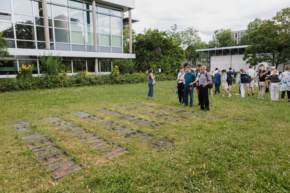 Auf einer Wiese sind die Buchstaben "Neben dem Kakaofeld" in den Boden eingelassen. Daneben steht eine Gruppe Menschen, die das Kunstwerk betrachtet.